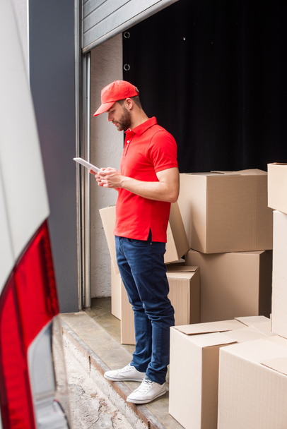 side view of delivery man in red uniform using digital tablet with cargo behind - Фото, изображение