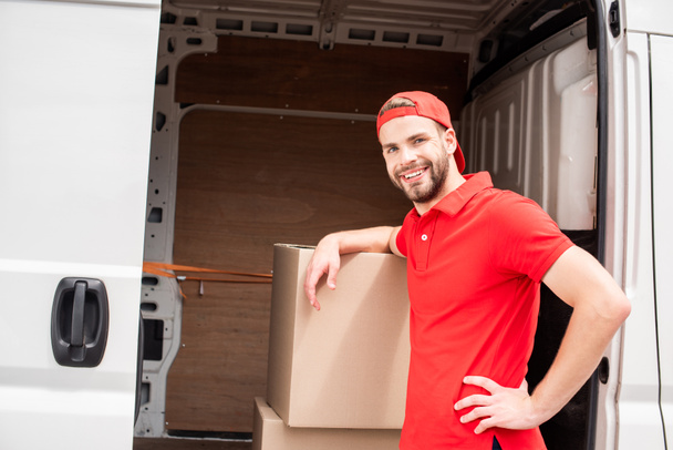 smiling young delivery man in uniform standing near van with cargo - 写真・画像