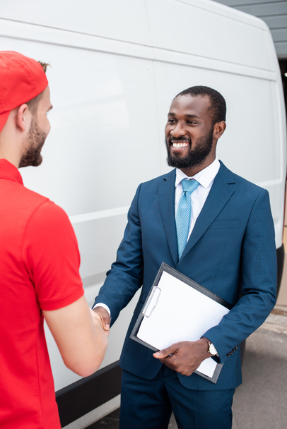 smiling delivery man and african american businessman shaking hands - Zdjęcie, obraz