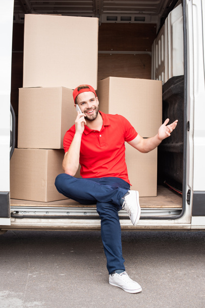 homem de entrega sorridente em uniforme vermelho falando no smartphone enquanto descansa em van com caixas de papelão
 - Foto, Imagem