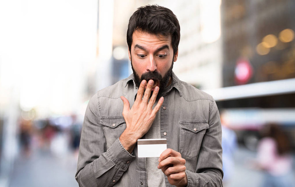 Удивлённый красавчик с бородой держит кредитку на улице
 - Фото, изображение