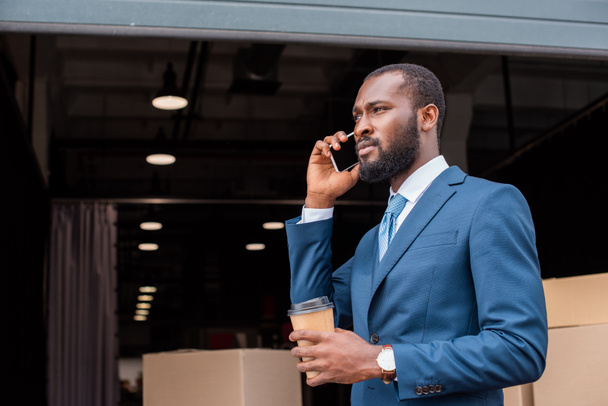 side view of african american businessman with coffee to go talking on smartphone - Фото, изображение