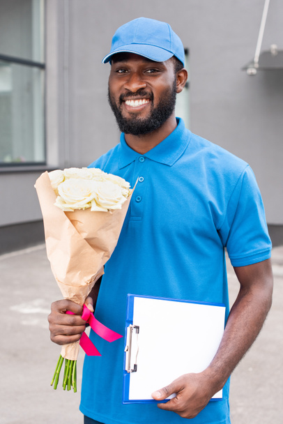 portrait of smiling african american delivery man with bouquet of flowers and cardboard in hands  - Photo, Image