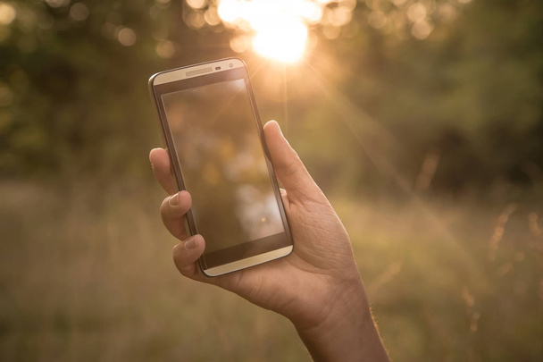 hand houden zwarte slimme telefoon, mobiele telefoon, mobiele over wazig beeld van groene bos achtergrond - Foto, afbeelding