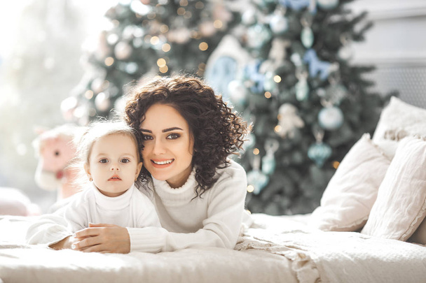 Young mother and her daughter having fun near the Christmas tree indoors - Photo, Image