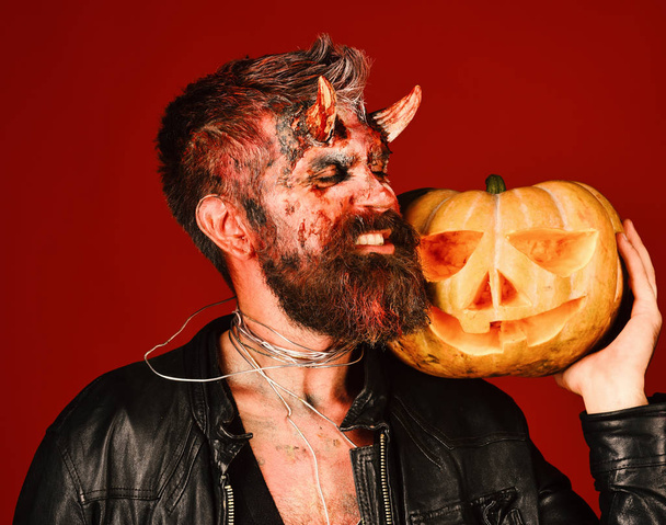 Man wearing scary makeup holds pumpkin on bloody red background. Devil or monster with October decorations. Demon with demonic face - Fotografie, Obrázek