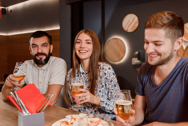 Two men and woman holding glasses of beer and sitting in pizzeria. - 写真・画像