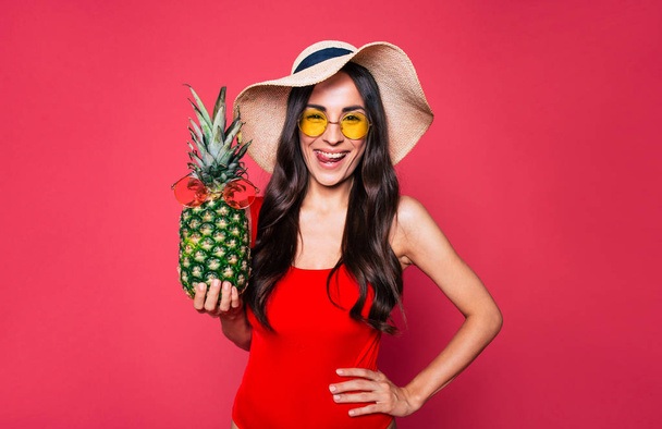 young brunette woman in red swimsuit, hat and glasses posing with pineapple on pink background  - Фото, зображення