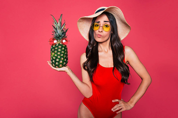 young brunette woman in red swimsuit, hat and glasses posing with pineapple on pink background  - Fotoğraf, Görsel