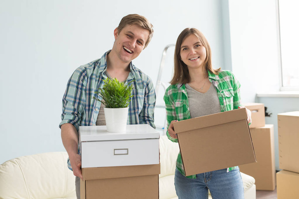 Furnuture, moving and people concept - beautiful happy couple moving in new flat and carry boxes and plant - Photo, Image