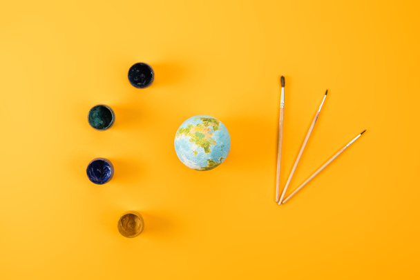 bovenaanzicht van earth globe met verf penselen en potten van gouache op geel - Foto, afbeelding