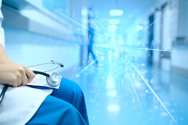 Женщина-врач со стетоскопом в руках сидит подавленная в больничном зале на фоне мигающего света
. - Фото, изображение