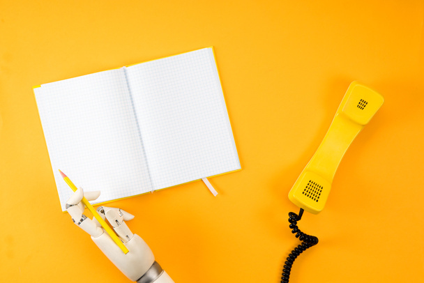 ビンテージ電話で黄色の卓上の空白のノートブックに書くロボットの手のショットをトリミング - 写真・画像