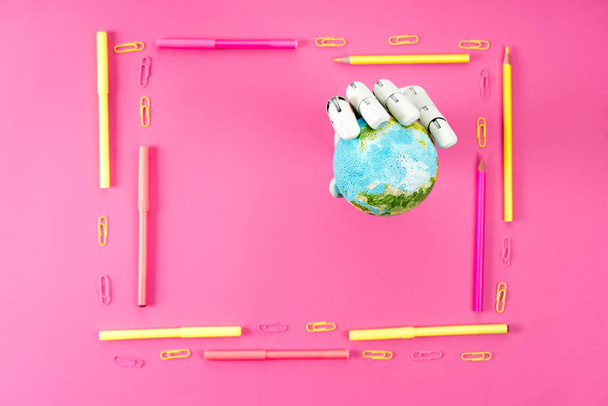 top view of robotic hand holding globe inside of frame made of pencils and markers on pink - Photo, Image