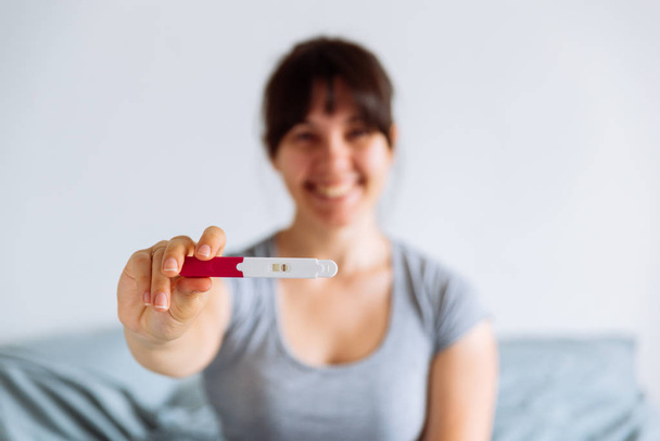 junge glückliche Frau, die auf dem Bett sitzt und auf einen positiven Schwangerschaftstest schaut. verschwommener Hintergrund - Foto, Bild