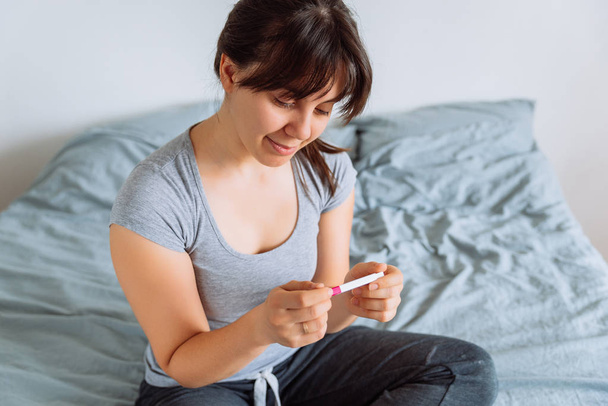mujer feliz joven sentada en la cama mirando en la prueba de embarazo positiva
 - Foto, imagen