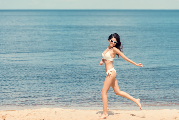cheerful girl in white bikini running on beach near the sea in summer - Фото, изображение