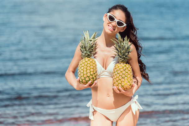 beautiful smiling woman in bikini and sunglasses holding fresh pineapples near the sea - Photo, Image