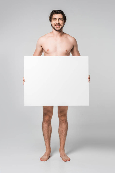 smiling handsome naked man holding blank banner on grey background - 写真・画像