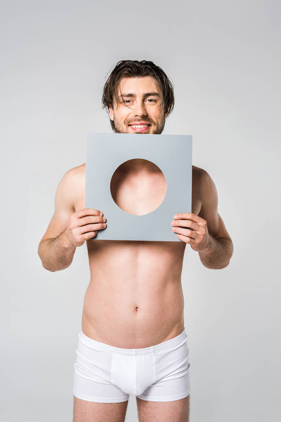 portrait of young smiling man in underwear holding circle paper figure isolated on grey - Photo, Image