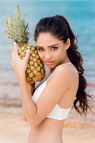 portrait of young brunette woman in white bikini holding sweet pineapple near the sea - Photo, image