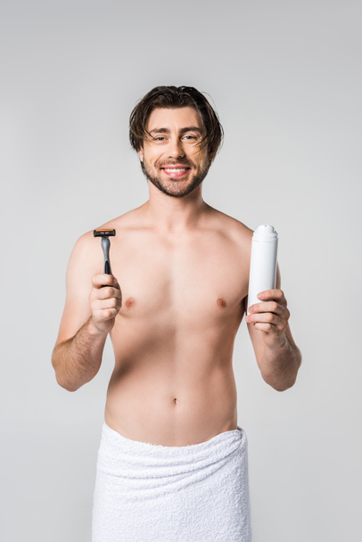portrait of smiling man in white towel with razor and shaving foam in hands isolated on grey - Photo, image