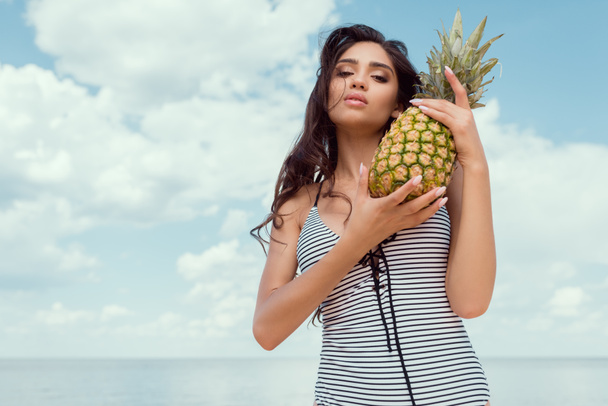 attractive brunette girl in swimsuit posing with pineapple near the sea - Fotografie, Obrázek