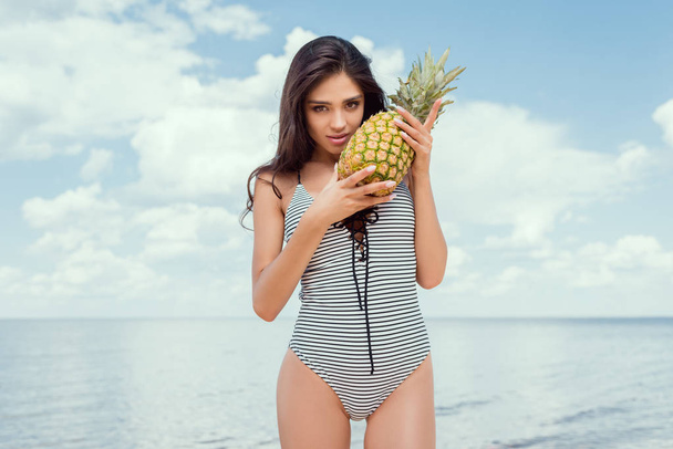 young brunette woman in swimsuit posing with pineapple near the sea in summer - Photo, Image