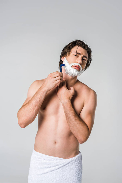 portrait of young man in white towel with shaving foam on face shaving beard isolated on grey - Фото, изображение