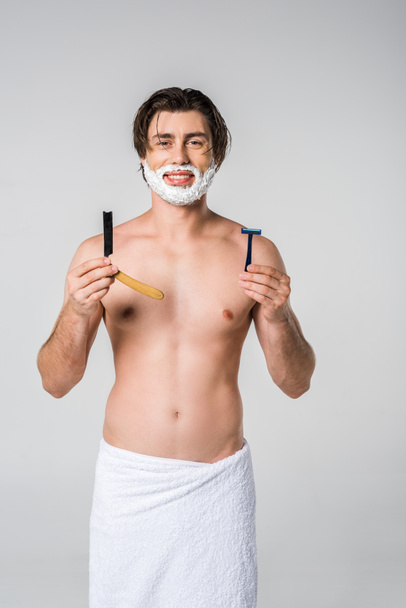 portrait of smiling man in white towel with shaving foam on face and razors in hands isolated on grey - Photo, image