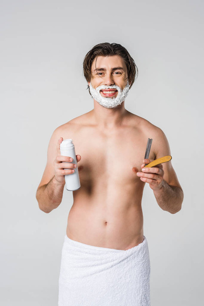 portrait of smiling man in white towel with shaving foam on face and straight razor in hand isolated on grey - Fotografie, Obrázek