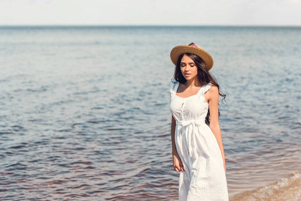 young woman in straw hat and white dress walking near the sea - Фото, зображення