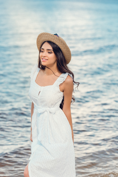 красивая счастливая девушка в соломенной шляпе и белом платье, гуляющая у моря
 - Фото, изображение