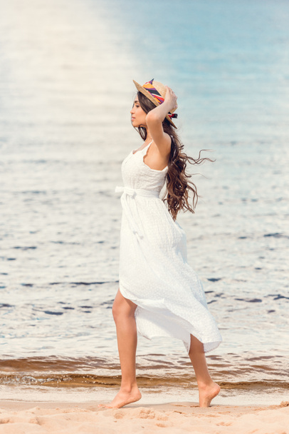 молодая женщина в соломенной шляпе и белом платье прогулка по песчаному пляжу
 - Фото, изображение