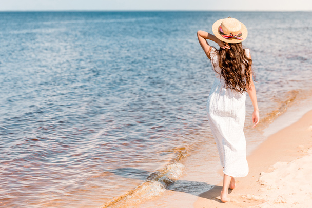 back view of woman in straw hat and white dress walking on beach near sea - Фото, изображение