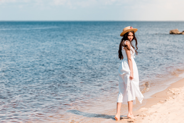 young woman in straw hat and white dress walking on beach - Фото, изображение
