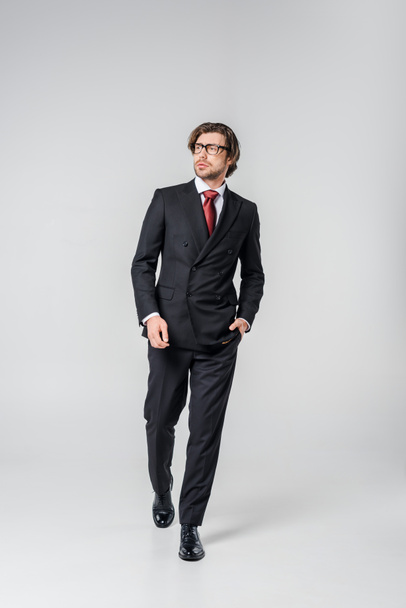 young businessman in stylish suit and eyeglasses walking on grey backdrop - 写真・画像