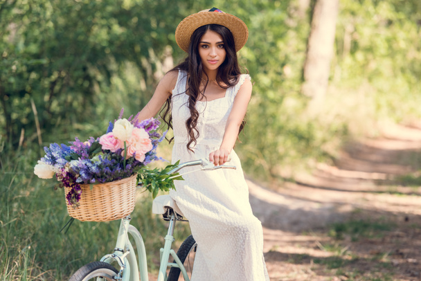 приваблива дівчина в солом'яному капелюсі сидить на велосипеді з квітами в плетеному кошику в парку
 - Фото, зображення