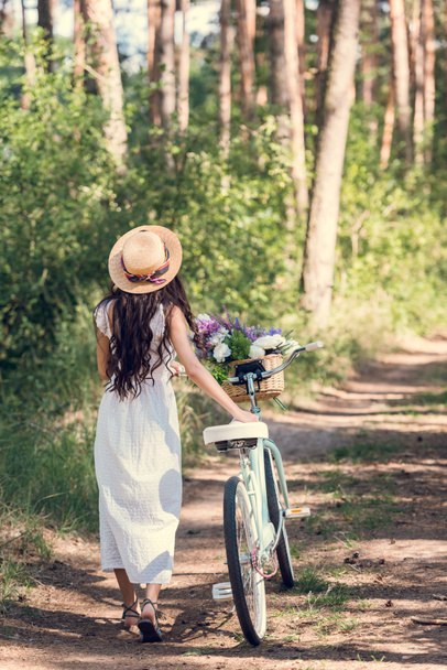 Achteraanzicht van meisje in stro hoed en witte jurk wandelen met fiets en bloemen in rieten mand - Foto, afbeelding