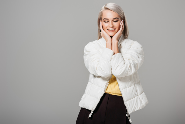 Lachende vrouw in witte jas poseren met handpalmen op gezicht geïsoleerd op een grijze achtergrond  - Foto, afbeelding