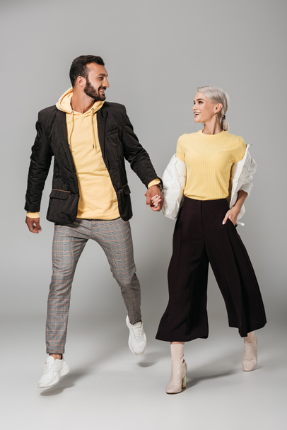 fashionable couple of models holding hands and looking at each other on grey background  - Фото, изображение