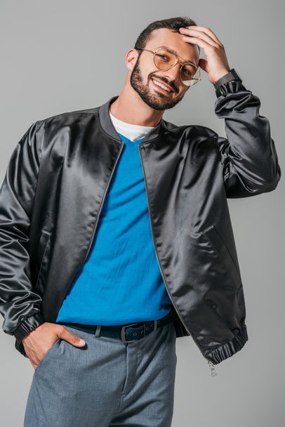 smiling male model in stylish black bomber posing with hand in pocket isolated on grey background  - Foto, imagen