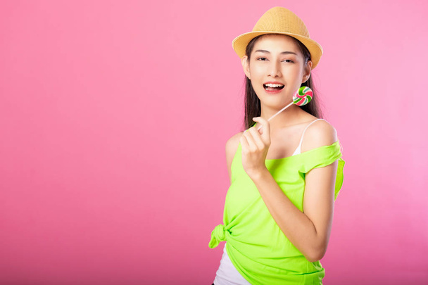 Portrait of a smiling attractive woman in summer outfit and hat holding lollipop and looking at camera isolated over pink background. - Photo, Image