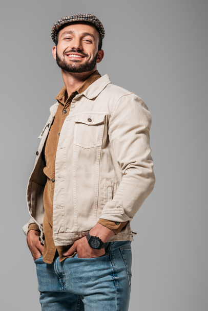 бородатый улыбающийся мужчина в осенней куртке и твидовом колпаке, позирующий с руками в карманах, изолированный на сером
 - Фото, изображение