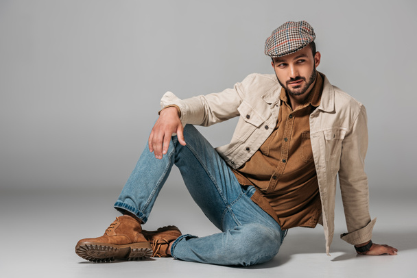 бородатий чоловік позує в шнуровій сорочці, джинсах і осінній куртці і твії шапці, на сірому
 - Фото, зображення