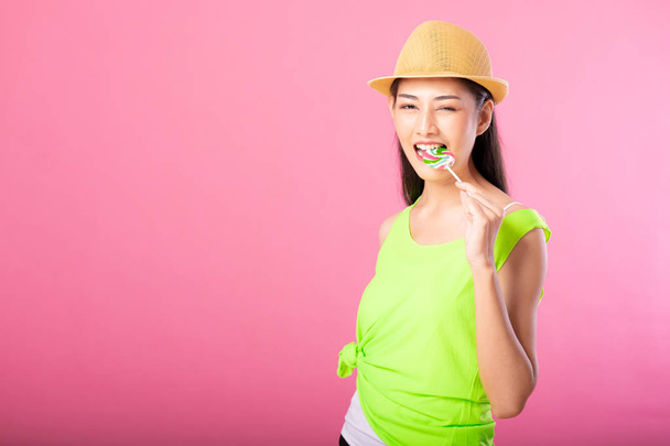 Portrait of a smiling attractive woman in summer outfit and hat biting lollipop and looking at camera isolated over pink background. - Фото, изображение