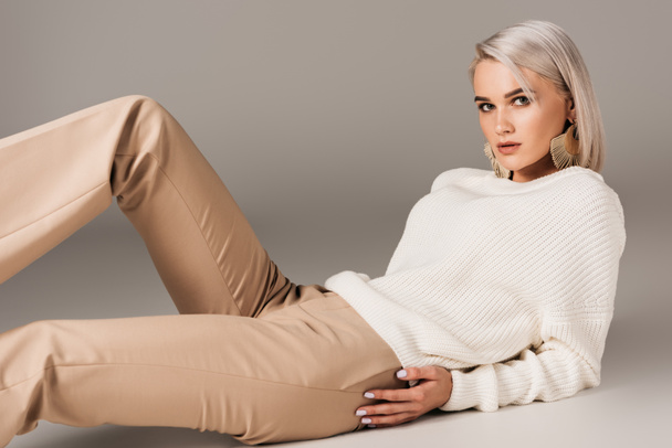 elegant blonde woman lying in white sweater and beige pants, on grey - Zdjęcie, obraz