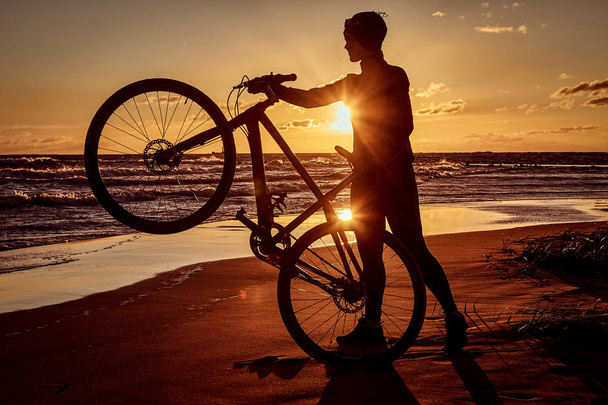 Силует жінки тримає велосипед на морському узбережжі на фоні заходу сонця
. - Фото, зображення