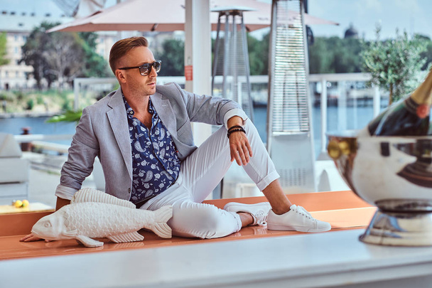 Tyylikäs mies pukeutunut moderni tyylikäs vaatteita istuu pöydällä kala veistos ulkona kahvilassa taustaa vasten kaupungin laituri
. - Valokuva, kuva