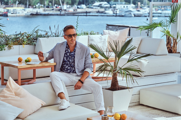 Fashionable successful man with stylish hair dressed in modern elegant clothes holds a cup of coffee while sitting on a sofa at outdoor cafe against the background of the city wharf. - Fotoğraf, Görsel